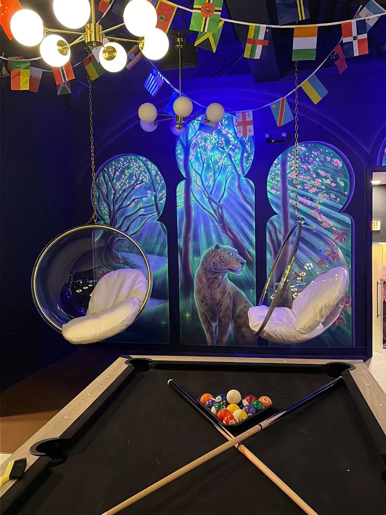 A pool table and hanging egg chairs beneath a glowing blossom-and-leopard mural